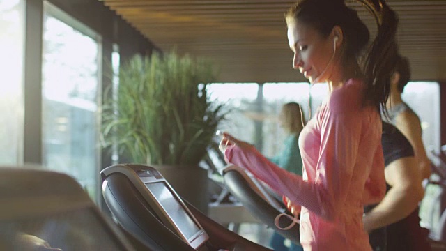 有吸引力的白人女孩在跑步机上在体育健身馆与电话和耳机。视频素材