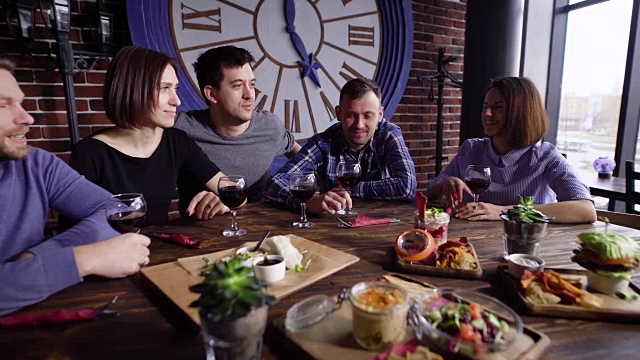 五个朋友拿着红酒坐在餐桌前，在餐厅里吃着不同的零食，开派对。男男女女都在靠窗的咖啡馆里消磨闲暇时光视频素材