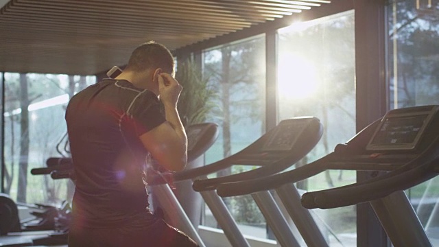一个健壮的运动员戴着耳机开始在体育馆里的跑步机上跑步。视频素材