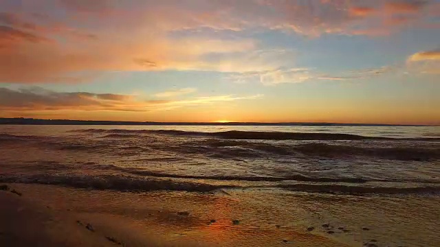 波浪在沙滩上翻滚。傍晚，太阳已经走出地平线。在远处，可以看到另一个大湖的河岸。美丽的红色天空。夏日黄昏后的日落。视频下载