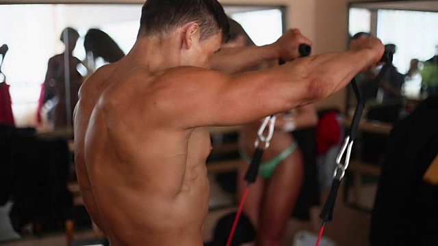 侧面，背部的男性健美运动员与裸露的躯干做运动与阻力带。背景中穿着比基尼的女人视频下载