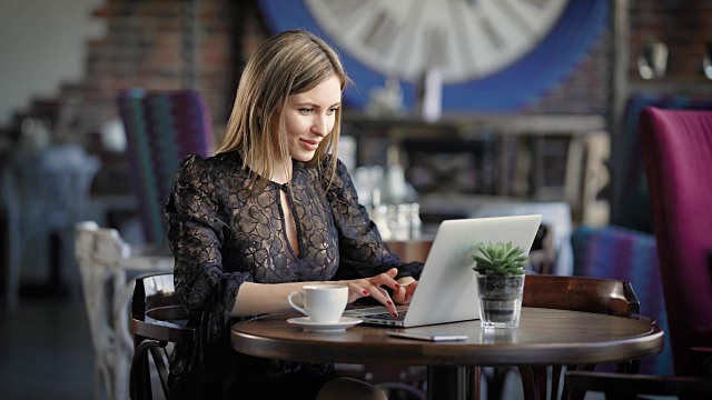 一个漂亮的女人在咖啡馆用笔记本电脑工作。一个快乐的女孩，在笔记本电脑的键盘上打印，她收到了好消息，并通过社交媒体分享它们视频下载