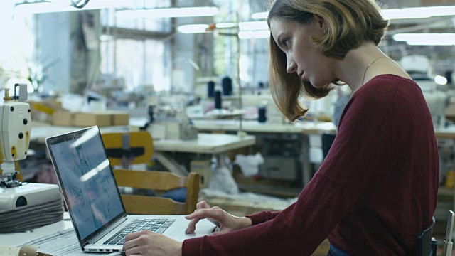 一名年轻女子站在一家服装厂里，正在使用智能手机，而工作人员在后台工作。视频素材