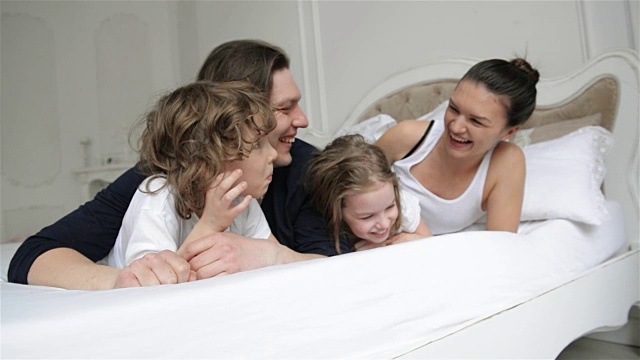 不安分和活跃的家庭一起在床上玩在家里的周末。年轻的父母带着迷人的微笑在卧室里和他们的儿子和女儿度过自由的时间视频下载