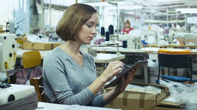 一名年轻女性正坐在服装厂的一张桌子旁，用手写笔在写字板上画素描。视频素材