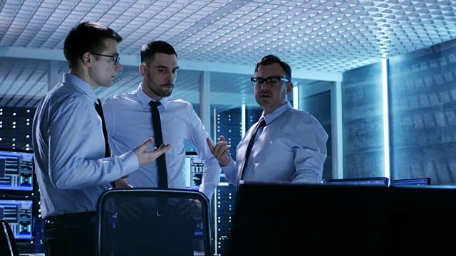 三位技术主持人在监控室进行积极的讨论。系统控制室充满了显示各种数据的工作显示器，并有服务器机架。视频素材
