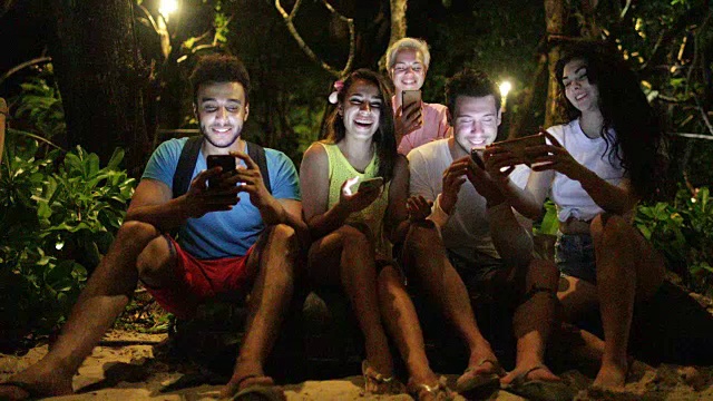 人们用手机在晚上公园的长椅上大笑，年轻的朋友在网上聊天视频素材