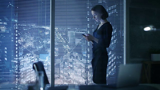 镜头聚焦在一个女商人透过百叶窗。她站在看大城市风景的窗口附近使用智能手机。视频素材