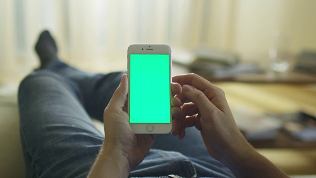一名男子躺在家里的沙发上，用绿色屏幕竖屏打电话。视频素材
