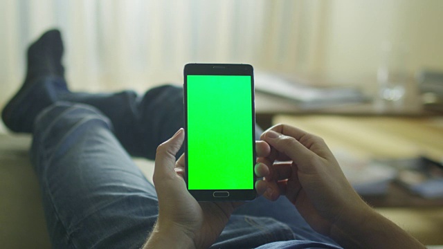 一名男子躺在家里的沙发上，在大腿上使用绿色屏幕的安卓手机。视频素材