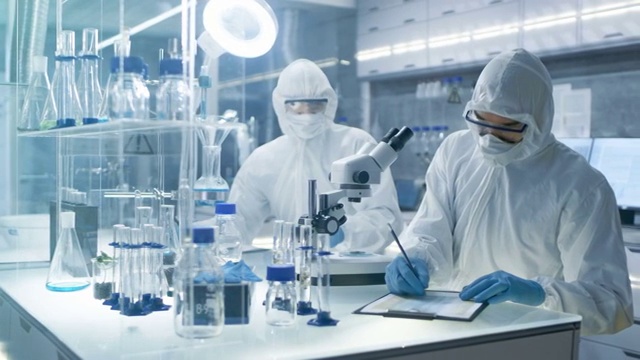 在一个安全的高水平实验室里，科学家们穿着工作服进行一项研究。化学家用钳子调整培养皿中的样品，然后在显微镜下检查它们，他的同事写下结果。视频购买
