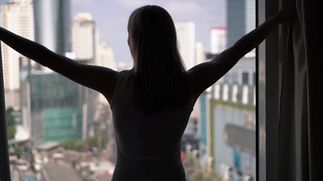 女人揭开窗帘向窗外望去的剪影。城市摩天大楼景观视频素材