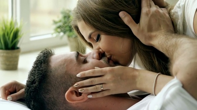 年轻美丽和相爱的夫妇在床上玩耍和亲吻在早上的特写。有魅力的男人亲吻和拥抱他的妻子在床上视频购买