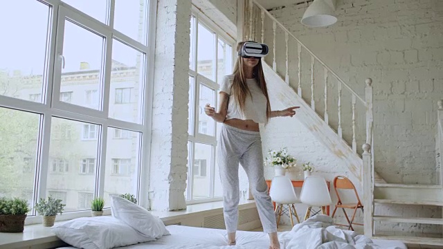 快乐微笑的年轻女子跳舞，同时获得体验使用360 VR头盔眼镜的虚拟现实在家床上视频下载