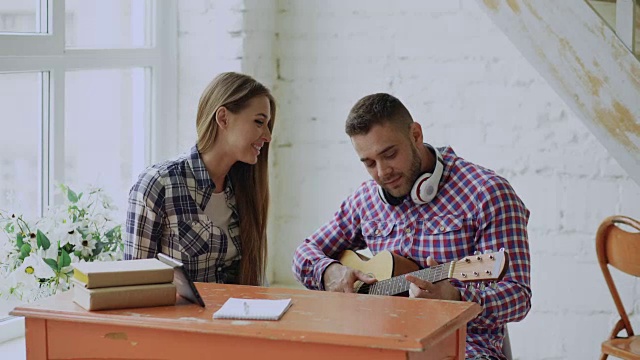 年轻的幸福和相爱的夫妇学习弹原声吉他，坐在家里的桌子上玩得很开心视频素材