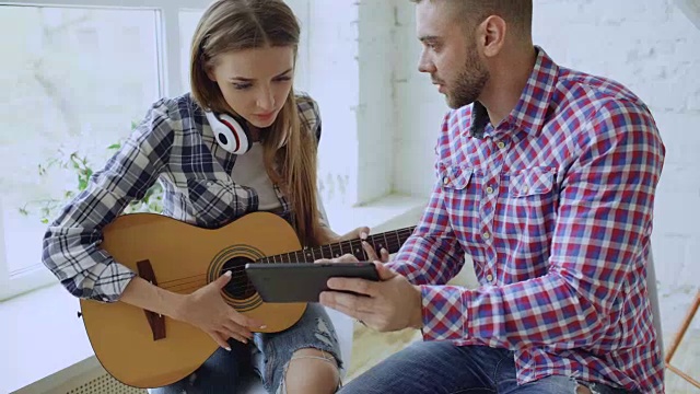 年轻的幸福和相爱的夫妇学习用平板电脑玩原声吉他，坐在家里的桌子上玩得很开心视频素材