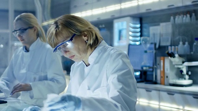 在大型现代实验室里在计算机上工作的女性和男性科学家。各种架子与烧杯，化学品和不同的技术设备可见。视频素材