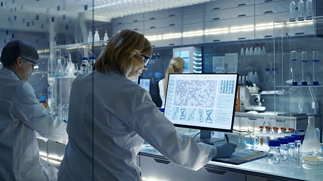 在大型现代实验室里在计算机上工作的女性和男性科学家。各种架子与烧杯，化学品和不同的技术设备可见。视频下载