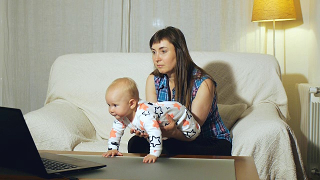 母亲和蓝眼睛的婴儿坐在白色沙发上看笔记本电脑屏幕上的视频视频下载