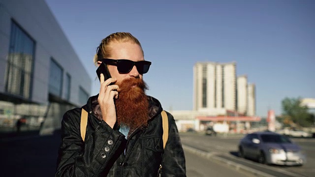 多莉拍到了一个戴着太阳镜、用智能手机聊天的年轻大胡子男人在城市街道上的照片视频素材