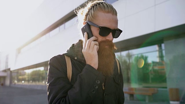 多莉拍到了一个戴着太阳镜、用智能手机聊天的年轻大胡子男人在城市街道上的照片视频素材