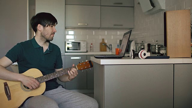 一个快乐的年轻人坐在厨房里用笔记本电脑学习弹吉他视频素材