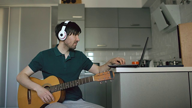 一个愉快的年轻人戴着耳机坐在厨房里用笔记本电脑学习弹吉他视频素材