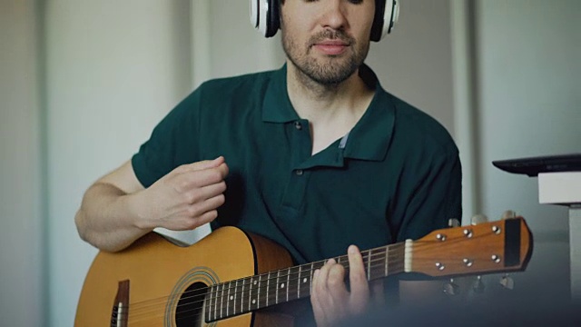 一个愉快的年轻人戴着耳机坐在厨房里用笔记本电脑学习弹吉他视频素材