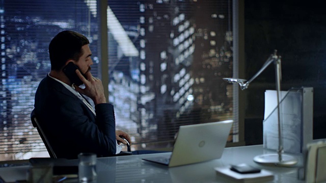 商人坐在办公桌前，在办公室里讲电话，可以看到大城市的风景。视频素材
