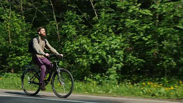 在一个晴朗的春日骑自行车散步。一名留着胡子的嬉皮士骑在路边，背景是绿色的森林和黄色的花朵视频素材