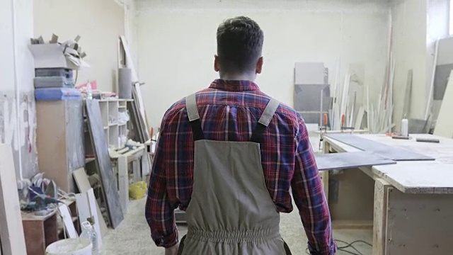 一个穿着工装裤的年轻木匠走在一家木工工厂的走廊上。视频素材