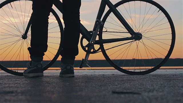 年轻英俊的潮人从地面上拿他的固定齿轮自行车在日落，户外视频素材