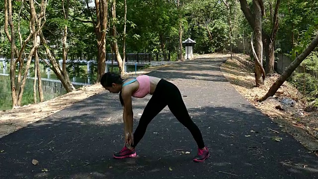 女性运动健身运动员正在为春天或夏天在森林小路上的户外慢跑做准备。健康年轻的亚洲妇女伸展跑步前或运动后在热带公园。视频下载