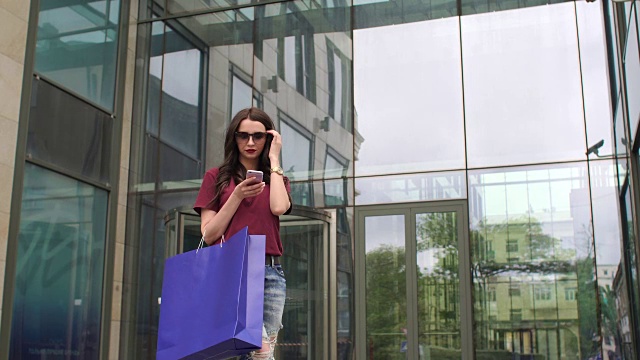 年轻女孩拿着手机和购物袋在购物中心附近。视频下载