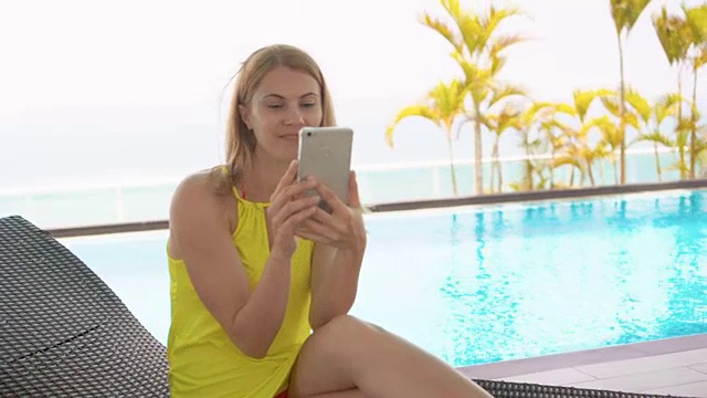 一名身穿黄色连衣裙的女子坐在泳池旁的日光浴床上用手机自拍视频下载