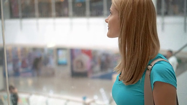 穿着绿松石t恤的年轻女子微笑着走下购物中心的电梯，向上看视频下载