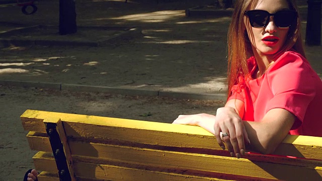 活泼迷人的女孩穿着红色套装，戴着黑色太阳镜坐在公园的长椅上视频素材