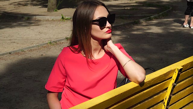 一位迷人的年轻女士穿着红色的衣服，戴着墨镜坐在公园的长椅上视频素材