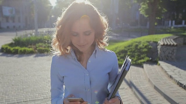 年轻漂亮的女商人，穿着蓝色衬衫，在城市的休息时间使用现代智能手机，职业女性雇主在外面用手机发短信视频素材