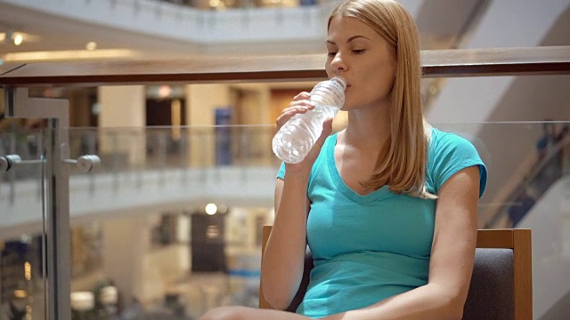 年轻美丽迷人的女人坐在购物中心的咖啡馆，用塑料瓶喝水视频素材