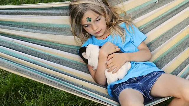金发小女孩坐在或躺在吊床上，抱着她的宠物豚鼠。视频素材