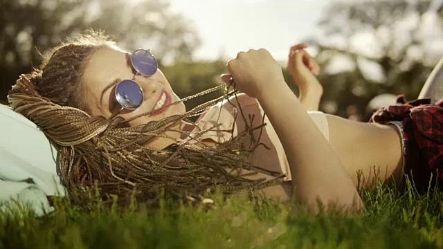 梳着辫子的时髦女孩在公园的草地上放松。夏天的生活方式肖像潮女人躺在草地上，享受美好的一天，戴着明亮的太阳镜。Slowmotion拍摄视频素材