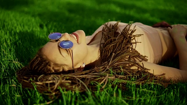 梳着辫子的时髦女孩在公园的草地上放松。夏天的生活方式肖像潮女人躺在草地上，享受美好的一天，戴着明亮的太阳镜。Slowmotion拍摄视频素材
