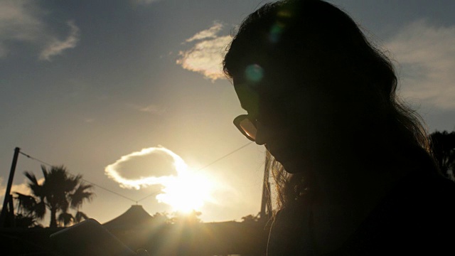 年轻女子用太阳镜平板电脑，对着太阳，云彩和棕榈树的剪影视频下载