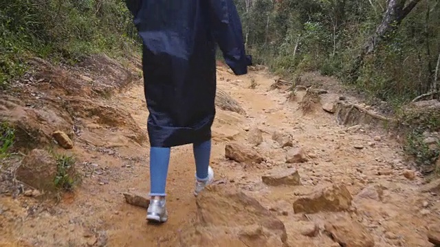 跟着一位穿着雨衣的女游客走在石山小道上。一个女人的脚徒步通过一个狭窄的峡谷。穿着运动鞋的女孩的腿走在崎岖的路上。低视角特写视频素材