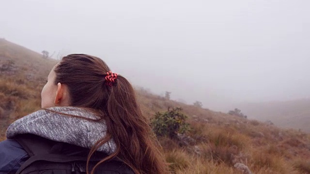 登山妇女在潮湿多雾的天气里穿着雨衣背着背包在山上行走。年轻女孩在旅行途中走山路。跟着女游客走在路上。后视图特写视频素材