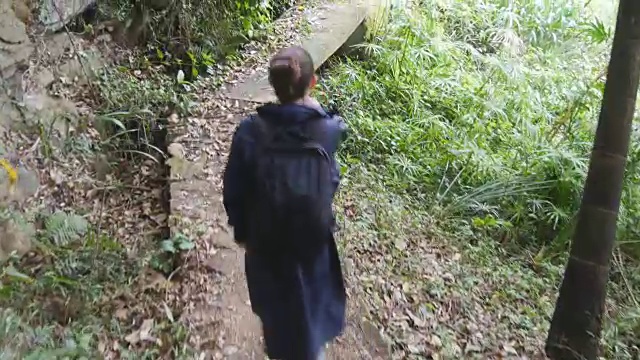 年轻的女孩在旅行中穿着雨衣在森林小径上。徒步女子背包行走在热带潮湿的森林。跟随女游客踩在丛林小径。后方后视图视频素材