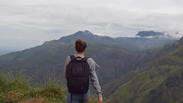 一名年轻女性徒步旅行者背着背包爬上山顶，举起双手。一名女游客站在美丽的峡谷边缘，胜利地伸出双臂。后方后视图视频素材