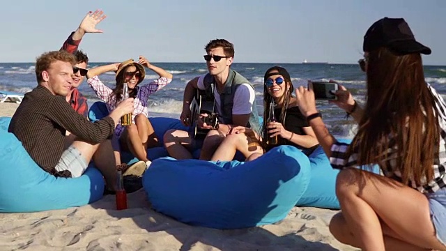 一个年轻的时髦女孩在一个夏天的晚上录制视频或拍摄一群朋友坐在沙滩上的安乐椅上弹吉他、唱歌的照片。Slowmotion拍摄视频下载