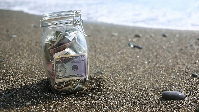一个装有纸币的玻璃罐子，以海浪为背景。在海边或海洋上积累休假的积蓄。海滩视频下载